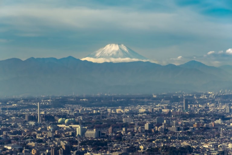 Snow-capped Mount Fuji towers over a hazy blue mountain range and a sprawling urban landscape under a soft sky.