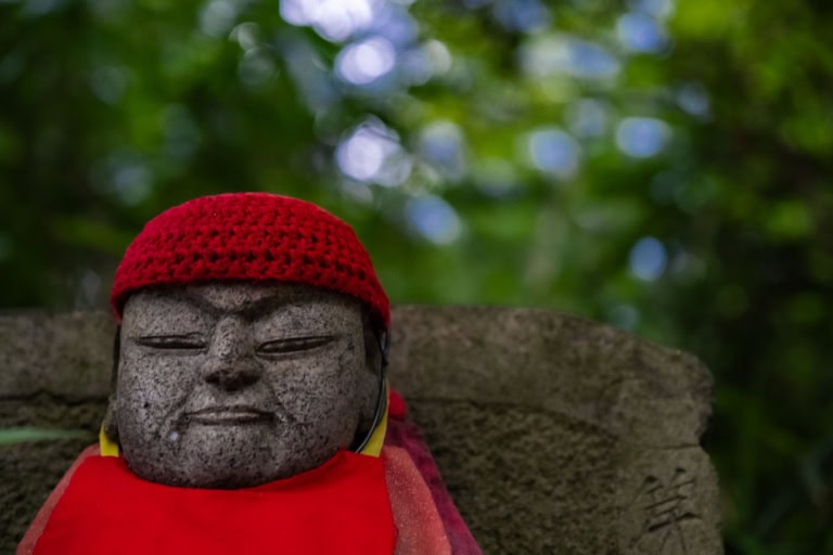 Jizo statue wearing a red knitted cap and bib at Takahata-Fudoson, set against a lush, blurred green background.