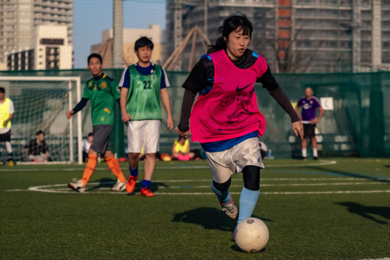 A woman in a pink bib dribbles a soccer ball on a turf field with other players and urban buildings in the background.