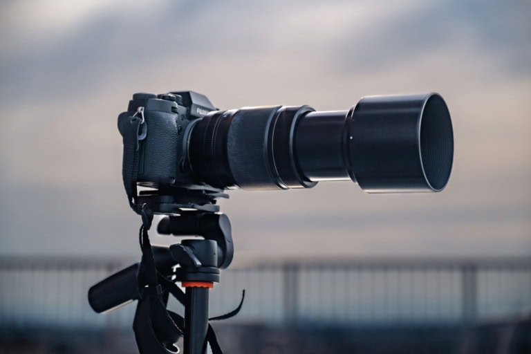 A Fujifilm camera on a tripod with a Fujinon XF 55-200mm lens, set against a soft, overcast horizon.