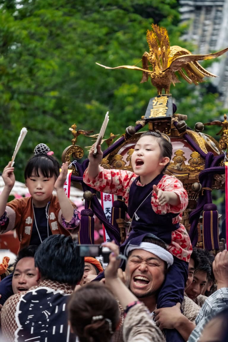 A child cheers on a man’s shoulders during the Sanja Festival 2018, with a golden mikoshi in the background at Asakusa.