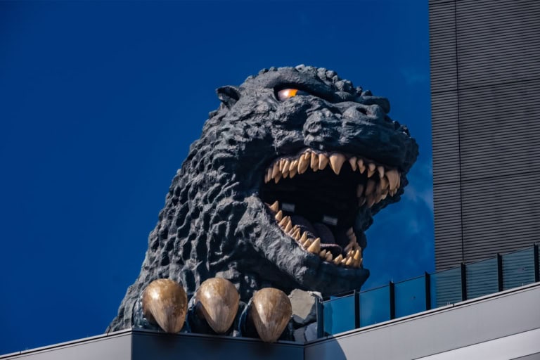 Godzilla head sculpture with sharp teeth and claws atop a Shinjuku building under a bright blue sky.
