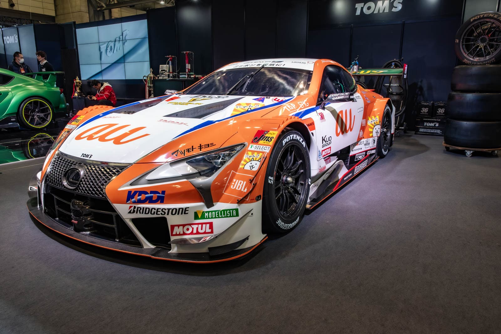 Au TOM'S LC 500 racecar at Tokyo Auto Salon 2020, displayed near TOM'S signage and stacked tires.