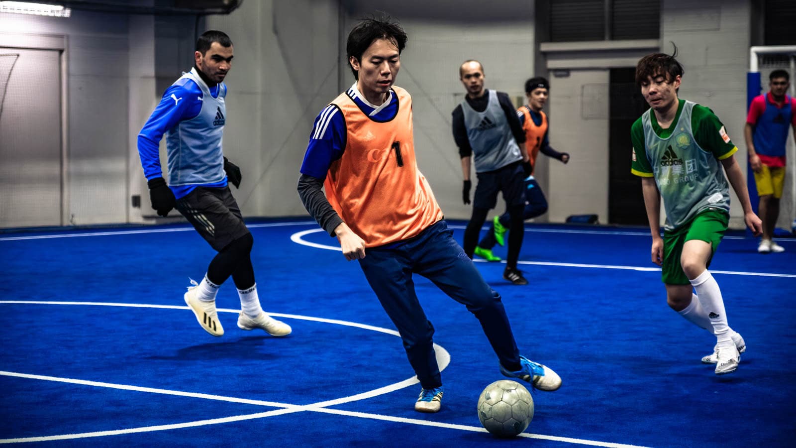 A man in an orange vest dribbles a soccer ball on a blue indoor court during a futsal match.
