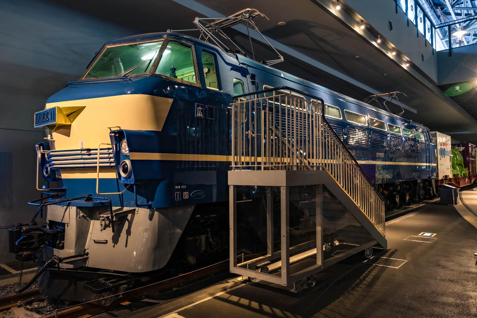Blue and cream EF66 electric locomotive on display indoors at a museum with a set of stairs beside it.