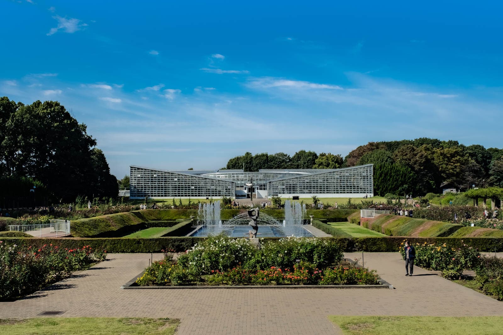 Jindai Botanical Garden featuring a central fountain and paths, with a large greenhouse and trees in the background under a blue sky.