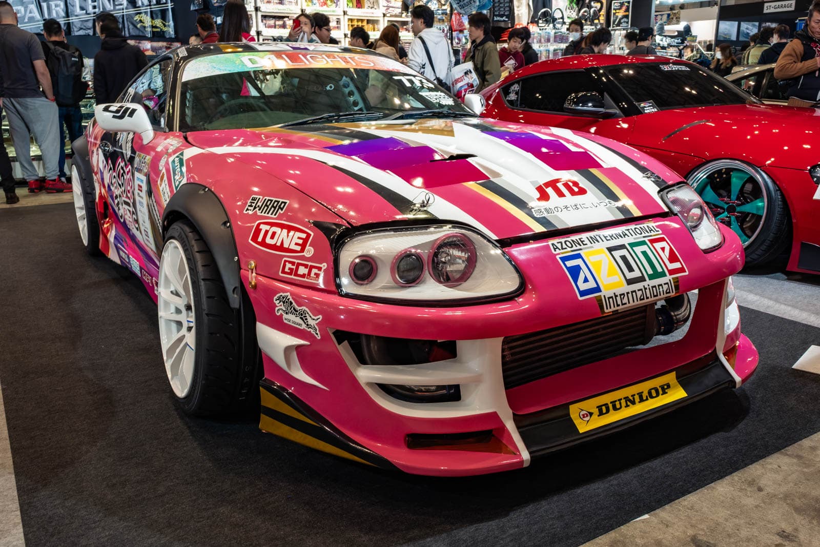 Pink Toyota Supra at Tokyo Auto Salon 2020, displayed near a red car and background crowds.