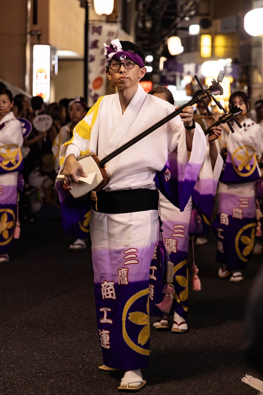 A musician in a white and purple kimono plays a shamisen during a nighttime street festival in Tokyo.