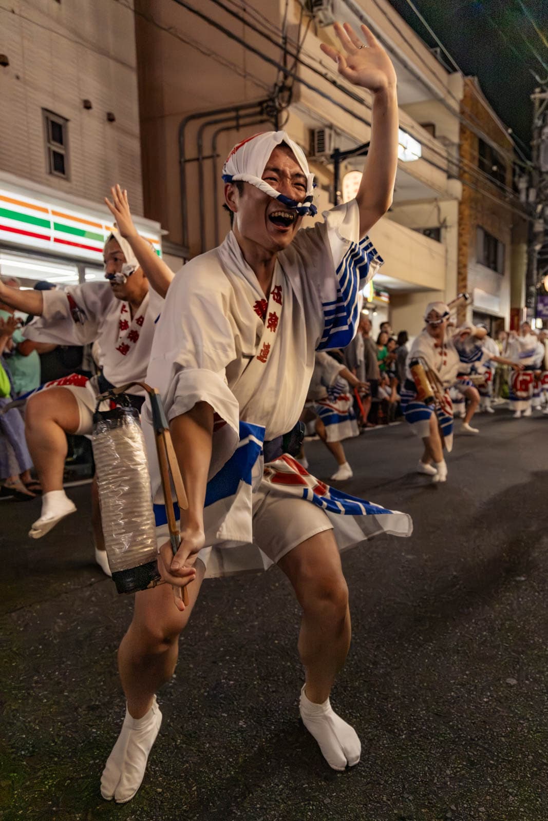 A smiling dancer in white and blue traditional attire raises an arm during a nighttime street festival in Tokyo.