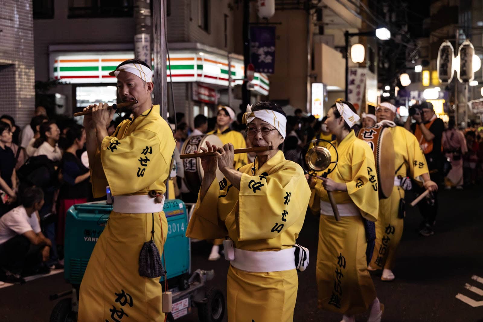 Performers in yellow happi coats and headbands play traditional flutes and drums during a nighttime street festival in Tokyo.