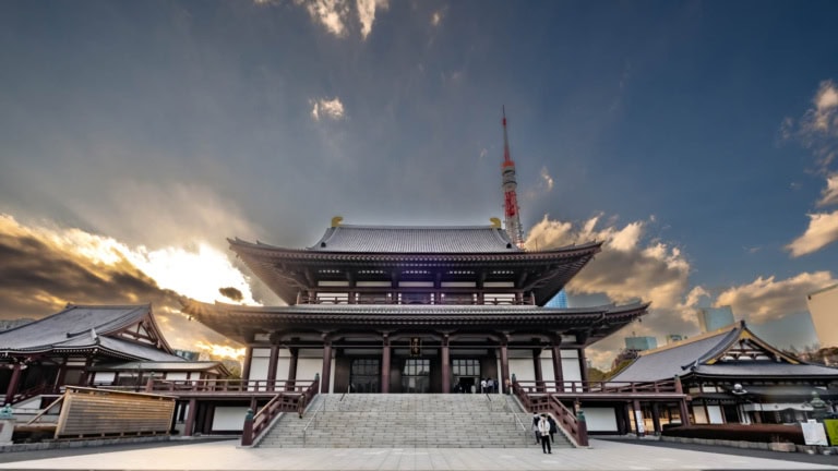 Zojoji Temple's main hall stands prominently above a wide staircase, viewed against a dramatic cloudy sky and Tokyo Tower.