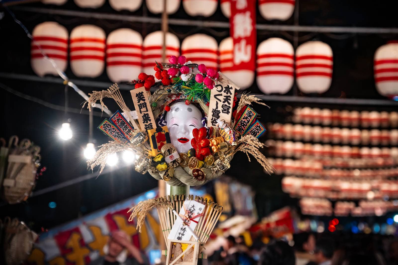 A kumade rake with a mask at Okunitama Shrine during the Tori-no-Ichi festival, under rows of red and white chochin lanterns.