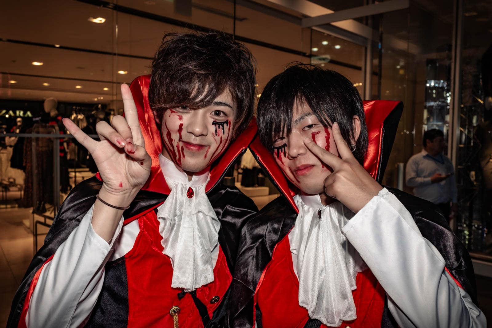 Two men in vampire costumes with high collars making peace signs during Halloween in Tokyo.