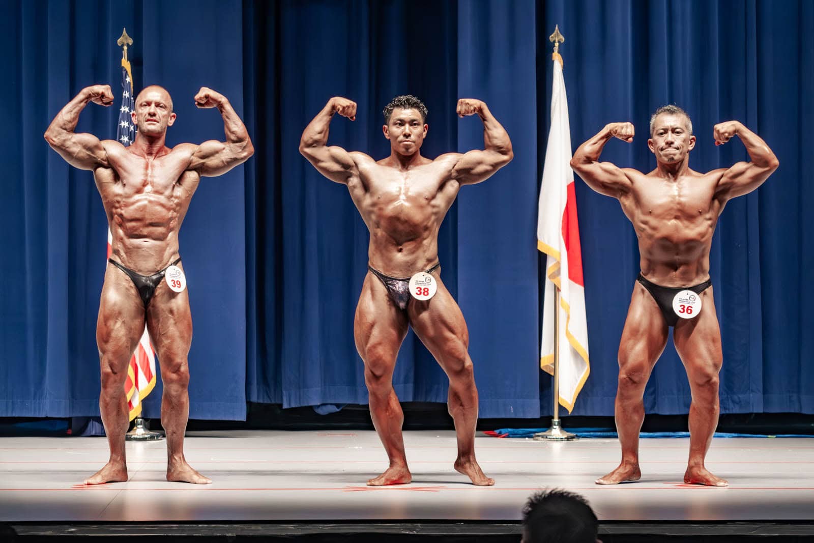 Three male bodybuilders double biceps pose before US and Japanese flags at the Taiyo Community Center at Yokota Air Base.
