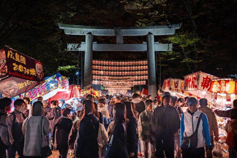A stone torii gate at Okunitama Shrine during a night festival, lined by Japanese street food stalls.