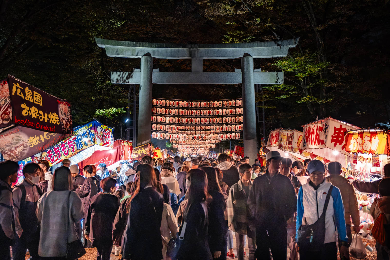 A stone torii gate at Okunitama Shrine during the Tori-no-Ichi festival, framed by night market food stalls.