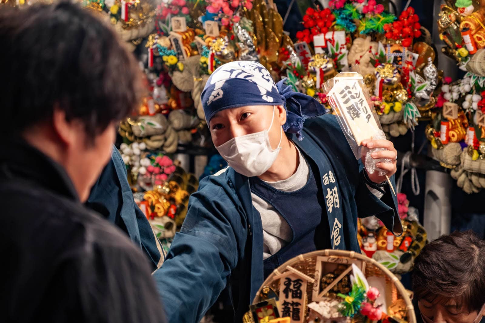 A sales staff member sells kumade rakes at Okunitama Shrine during the Tori-no-Ichi festival, with festival stalls behind.