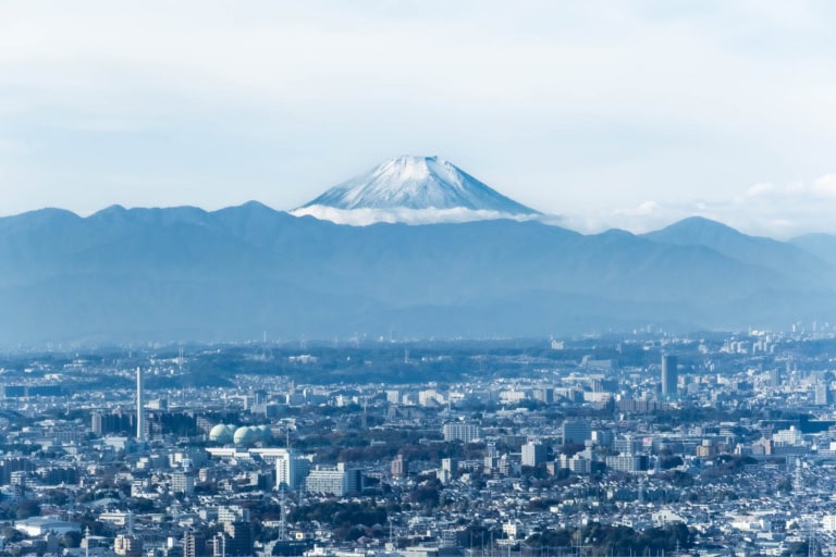 Snow-capped Mount Fuji rises above a mountain range and the Tokyo urban landscape on a cloud day