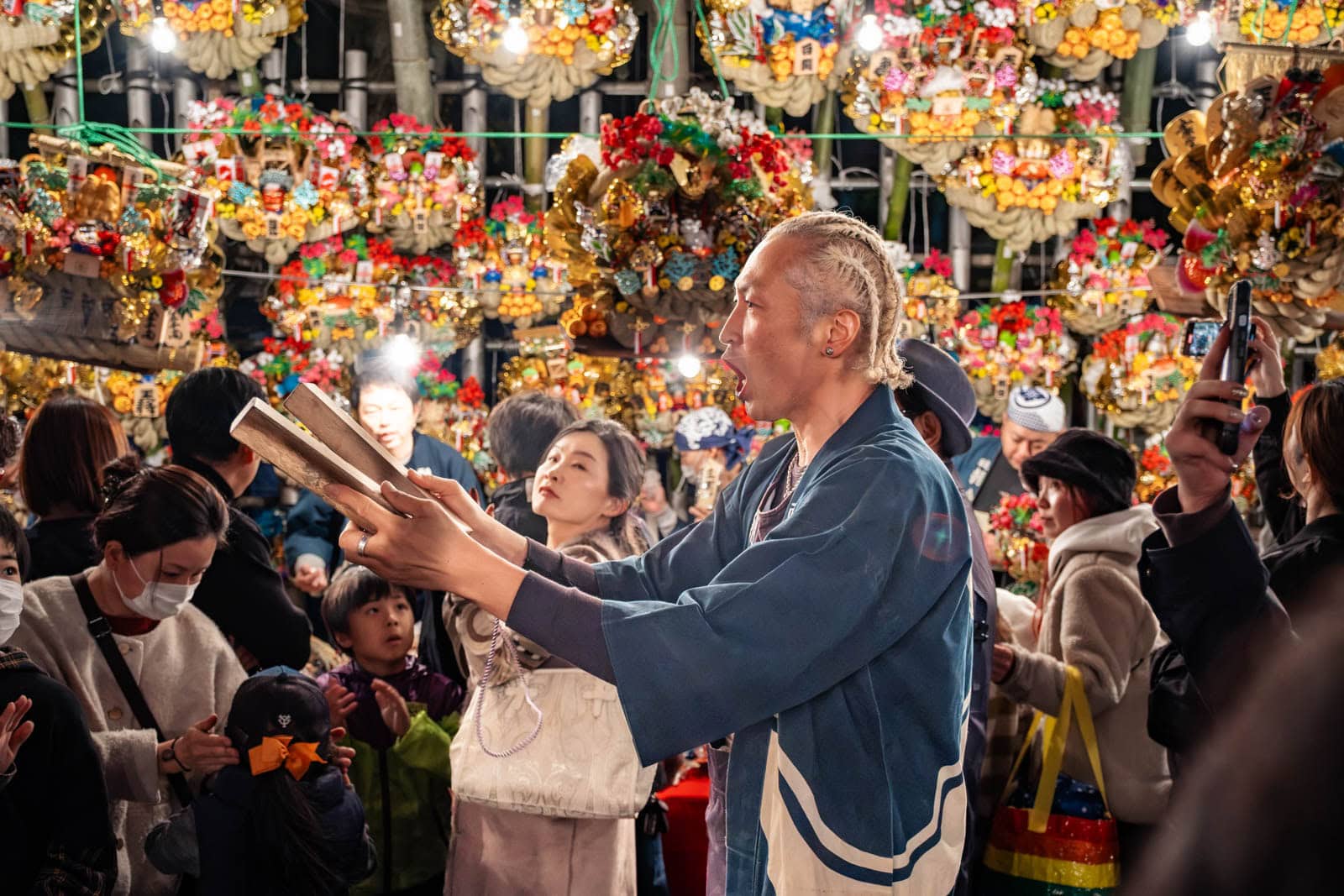 A man in a blue happi coat claps wooden clappers during the Tori-no-Ichi festival, surrounded by kumade rakes at a stall.